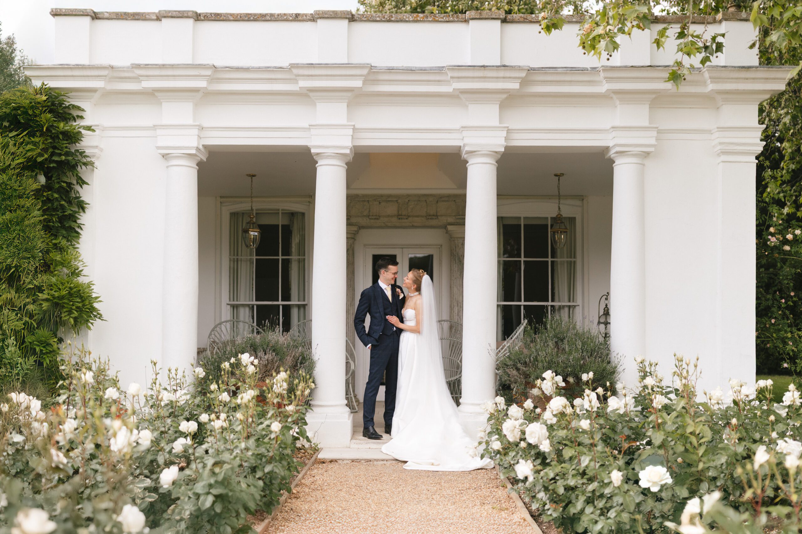 Bride and groom standing together outside Avington Park, framed by white columns and gardens, captured in a natural documentary wedding photography style.