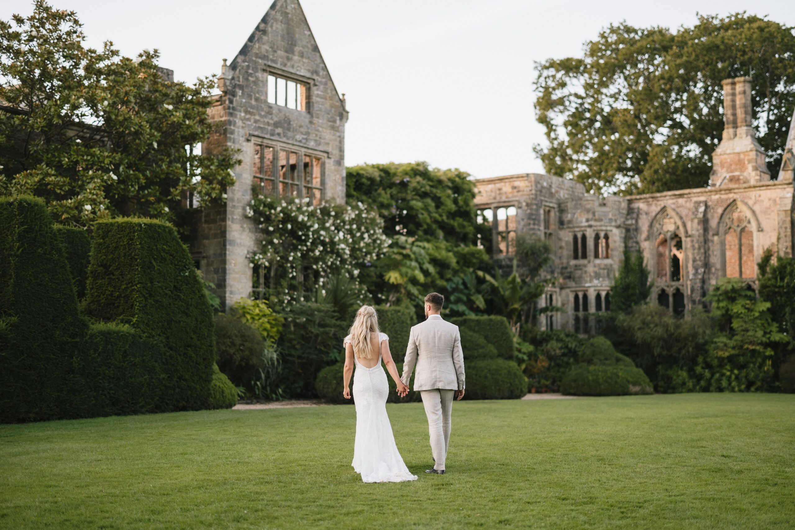Bride and groom walking hand in hand across the gardens at Nymans National Trust, with the romantic ruins and lush greenery creating a timeless, elegant backdrop.